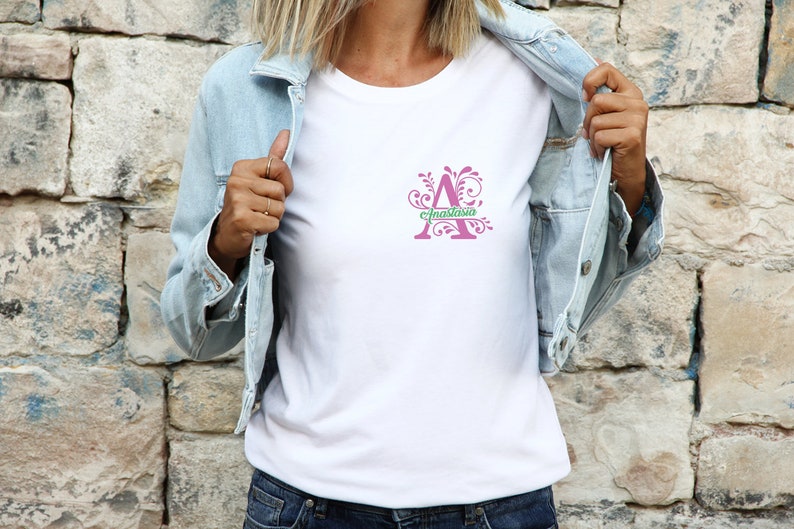 May include: A white t-shirt featuring a pink letter A with a green floral design. The letter A has the name "Anastasia" written across it.