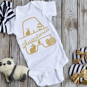 K&ouml;nnte beinhalten: Ein wei&szlig;er Baby-Strampler mit einer goldenen Grafik des Buchstabens A mit Hasen, K&uuml;ken und Ostereiern. Der Strampler ist mit dem Namen Anastasia personalisiert. Der Strampler liegt auf einem Holzgrund mit einem schwarz-wei&szlig;en Stirnband, einem Pandab&auml;rchen und schwarz-wei&szlig;en Schuhen mit goldenen Quasten.