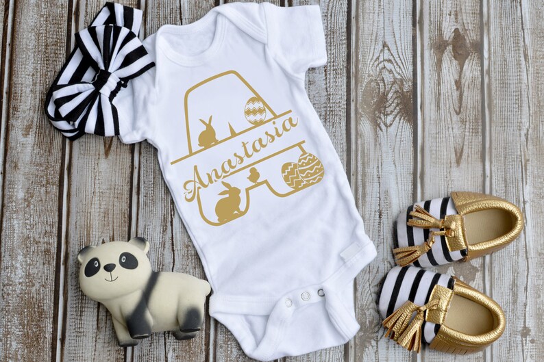 K&ouml;nnte beinhalten: Ein wei&szlig;er Baby-Strampler mit einer goldenen Grafik des Buchstabens A mit Hasen, K&uuml;ken und Ostereiern. Der Strampler ist mit dem Namen Anastasia personalisiert. Der Strampler liegt auf einem Holzgrund mit einem schwarz-wei&szlig;en Stirnband, einem Pandab&auml;rchen und schwarz-wei&szlig;en Schuhen mit goldenen Quasten.