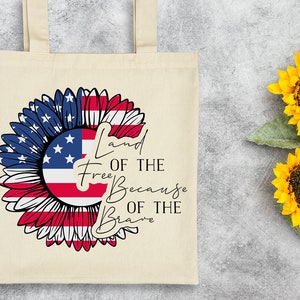 Puede incluir: Una bolsa de tela blanca con un dise&ntilde;o de girasol rojo, blanco y azul. El girasol est&aacute; hecho con la bandera estadounidense. El texto "Land of the Free Because of the Brave" est&aacute; impreso en la bolsa.