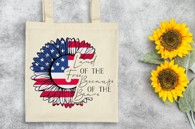 Puede incluir: Una bolsa de tela blanca con un dise&ntilde;o de girasol rojo, blanco y azul. El girasol est&aacute; hecho con la bandera estadounidense. El texto "Land of the Free Because of the Brave" est&aacute; impreso en la bolsa.