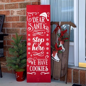 May include: A red wooden sign with white text that reads "Dear Santa stop here we have cookies". The sign is decorated with snowflakes and a Santa hat. The sign is leaning against a brick wall and there is a small Christmas tree in a red pot next to it.