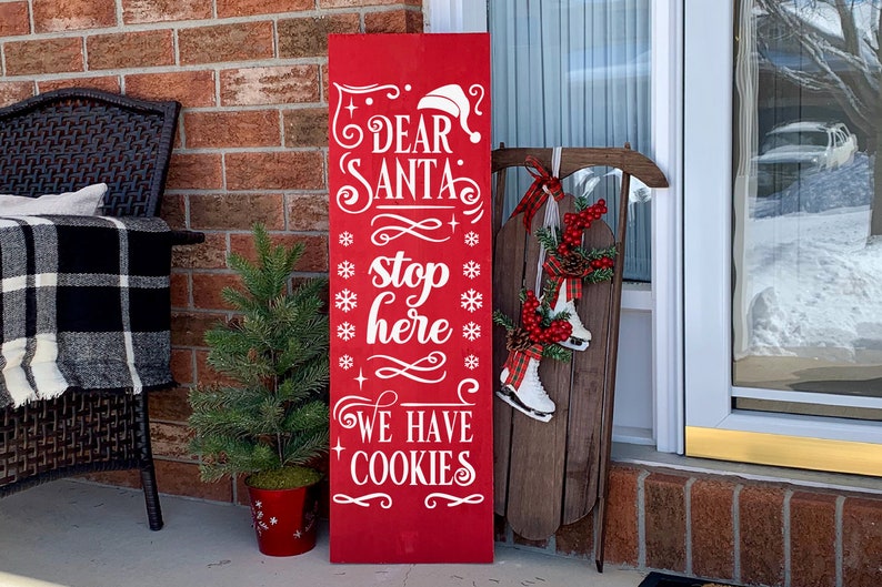 May include: A red wooden sign with white text that reads "Dear Santa stop here we have cookies". The sign is decorated with snowflakes and a Santa hat. The sign is leaning against a brick wall and there is a small Christmas tree in a red pot next to it.