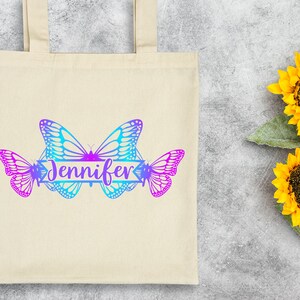 Op de afbeelding: Een witte canvas tote bag met een ontwerp van drie vlinders in tinten blauw en paars. De vlinders zijn in een rij gerangschikt met een banner waarop "Jennifer" in een cursief schrift staat.