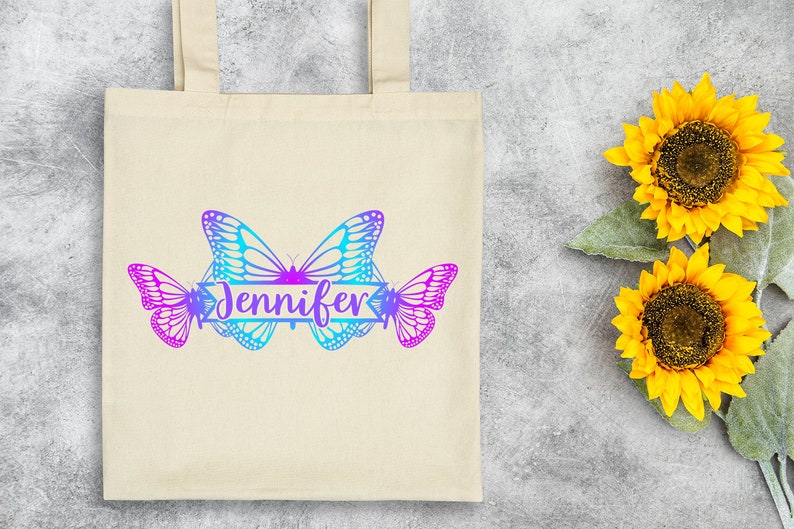 Op de afbeelding: Een witte canvas tote bag met een ontwerp van drie vlinders in tinten blauw en paars. De vlinders zijn in een rij gerangschikt met een banner waarop "Jennifer" in een cursief schrift staat.