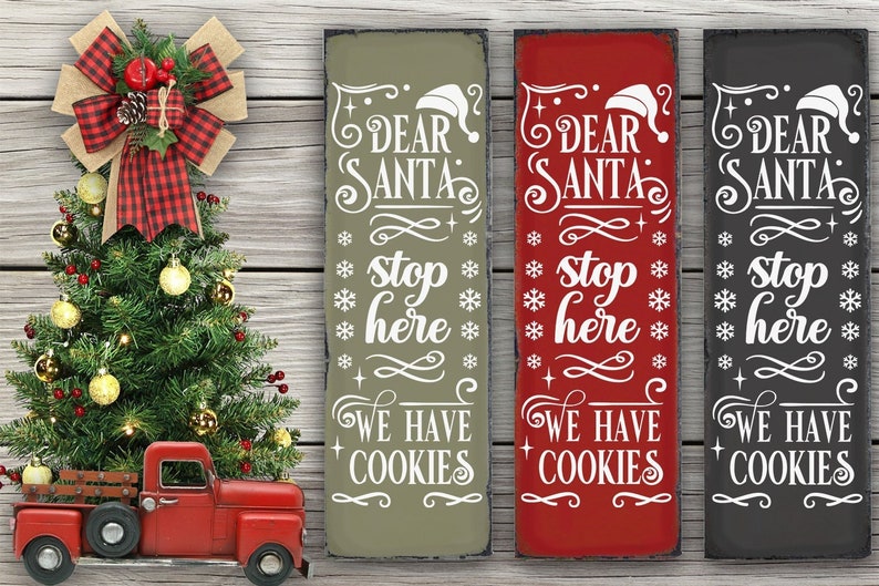 May include: Three wooden signs with the text "Dear Santa Stop Here We Have Cookies" in white lettering on a green, red, and black background. The signs are decorated with snowflakes and a Santa hat.