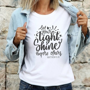 May include: White t-shirt featuring the phrase "let your light shine before others" in black script. The shirt is worn with a light blue denim jacket and blue jeans. The text includes the Bible verse Matthew 5:16.
