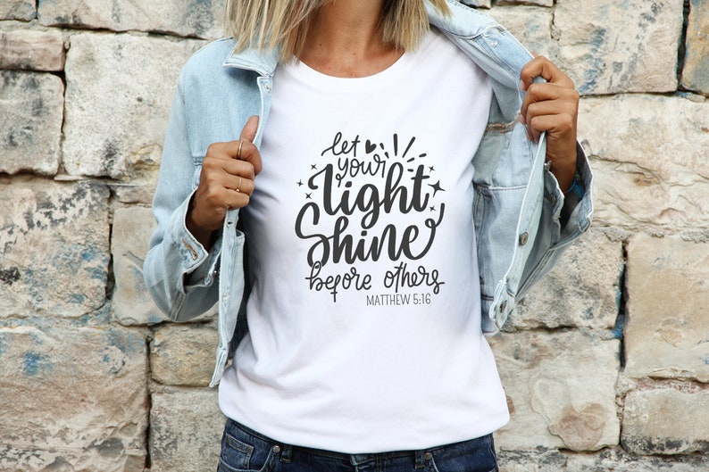 May include: White t-shirt featuring the phrase "let your light shine before others" in black script. The shirt is worn with a light blue denim jacket and blue jeans. The text includes the Bible verse Matthew 5:16.