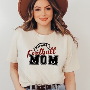 May include: A cream-colored short-sleeve t-shirt featuring a graphic print with a black football icon above the word Football in dark red script and the word MOM in large black block letters with a white outline on the front of the garment.