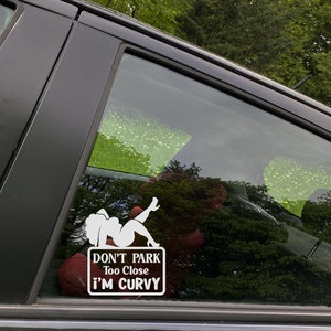 Puede incluir: Una pegatina de vinilo blanco en la ventana de un coche negro. La pegatina presenta a una mujer de dibujos animados con el texto "No aparques demasiado cerca, soy curvil&iacute;nea".