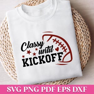 May include: White t-shirt with the text "Classy until Kickoff" in black and red, featuring a football graphic. The shirt is displayed on a woven, circular surface.