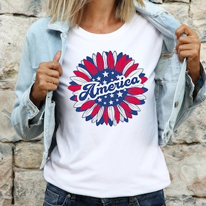 Puede incluir: Camiseta blanca con un dise&ntilde;o de girasol rojo, blanco y azul. El girasol tiene estrellas en el centro y la palabra "America" escrita en azul, con letra cursiva.