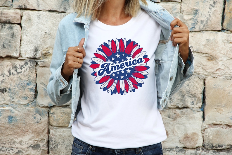 Puede incluir: Camiseta blanca con un dise&ntilde;o de girasol rojo, blanco y azul. El girasol tiene estrellas en el centro y la palabra "America" escrita en azul, con letra cursiva.