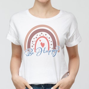 Pode incluir: Camiseta branca com um design de arco-&iacute;ris em tons de rosa e marrom. O arco-&iacute;ris &eacute; decorado com estrelas e dois cora&ccedil;&otilde;es vermelhos. O texto "Be Happy" est&aacute; escrito em azul cursivo abaixo do arco-&iacute;ris.