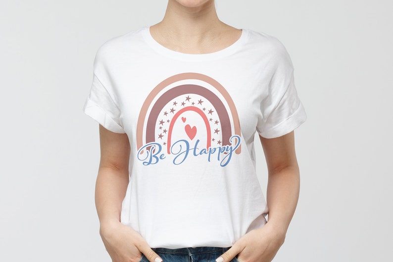 Pode incluir: Camiseta branca com um design de arco-&iacute;ris em tons de rosa e marrom. O arco-&iacute;ris &eacute; decorado com estrelas e dois cora&ccedil;&otilde;es vermelhos. O texto "Be Happy" est&aacute; escrito em azul cursivo abaixo do arco-&iacute;ris.