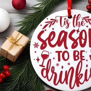 May include: White round Christmas ornament with red text that reads "Tis the season to be drinkin". The ornament is surrounded by green pine branches and red berries.