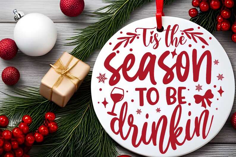 May include: White round Christmas ornament with red text that reads "Tis the season to be drinkin". The ornament is surrounded by green pine branches and red berries.