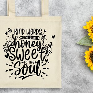 May include: A white tote bag with black text that reads "Kind words are like honey sweet to the soul". The text is in a handwritten font and is surrounded by small hearts and bees. The bag is sitting on a gray surface with two yellow sunflowers.