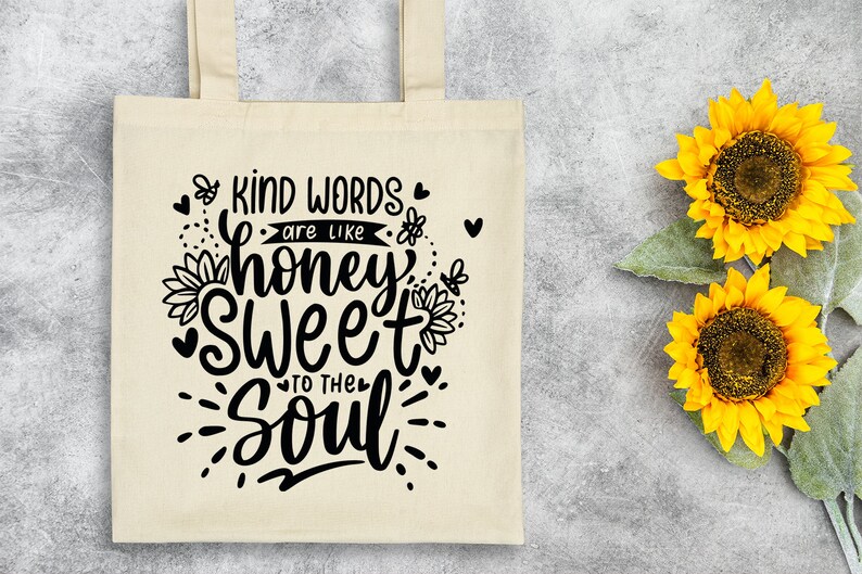 May include: A white tote bag with black text that reads "Kind words are like honey sweet to the soul". The text is in a handwritten font and is surrounded by small hearts and bees. The bag is sitting on a gray surface with two yellow sunflowers.