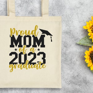 May include: A white canvas tote bag with black text that reads "Proud Mom of a 2023 Graduate" with a black graduation cap above the word "Mom". The text is in a bold, blocky font. The bag has a gold star on either side of the year 2023. The text "Graduate" is in a cursive font. The bag is on a gray background with two yellow sunflowers.