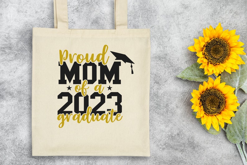 May include: A white canvas tote bag with black text that reads "Proud Mom of a 2023 Graduate" with a black graduation cap above the word "Mom". The text is in a bold, blocky font. The bag has a gold star on either side of the year 2023. The text "Graduate" is in a cursive font. The bag is on a gray background with two yellow sunflowers.