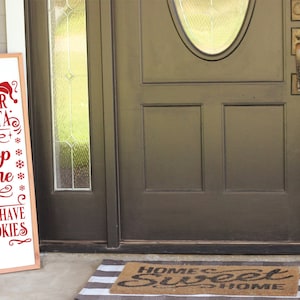 May include: A wooden sign with the text "Dear Santa Stop Here We Have Cookies" in red lettering. The sign is leaning against a brown door with a glass window.