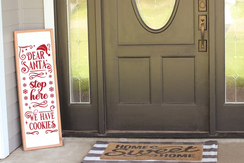 May include: A wooden sign with the text "Dear Santa Stop Here We Have Cookies" in red lettering. The sign is leaning against a brown door with a glass window.