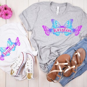 Op de afbeelding: Twee witte T-shirts met een vlinderontwerp en de namen "Sophie" en "Kristin" erop gedrukt. De shirts liggen op een wit houten oppervlak met een paar witte sneakers, een paar bruine sandalen en een blauwe jeans.