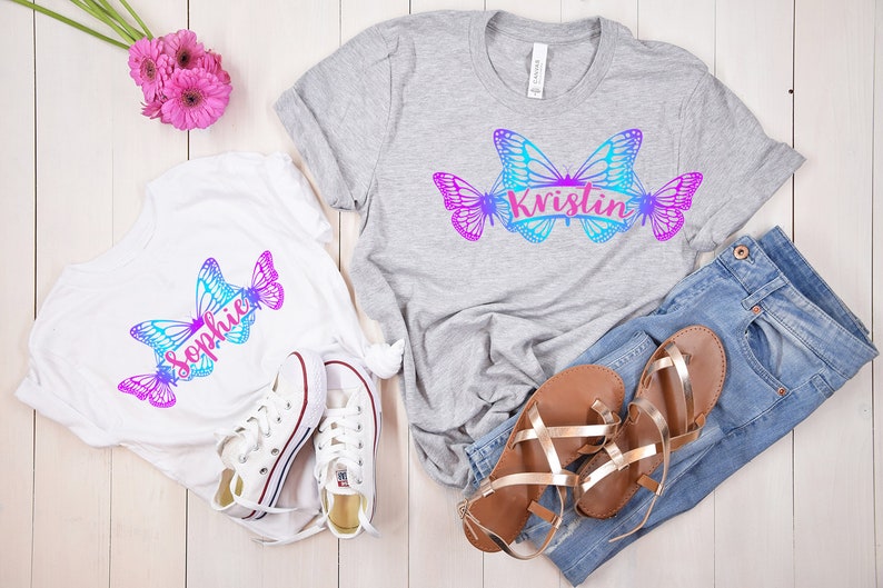 Op de afbeelding: Twee witte T-shirts met een vlinderontwerp en de namen "Sophie" en "Kristin" erop gedrukt. De shirts liggen op een wit houten oppervlak met een paar witte sneakers, een paar bruine sandalen en een blauwe jeans.