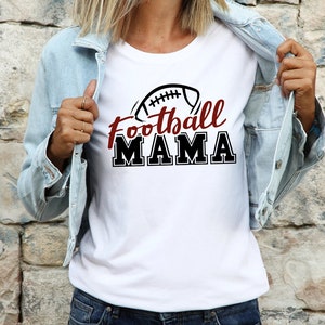May include: A white t-shirt featuring a graphic of an American football above the text Football MAMA in a combination of red script and black block lettering, worn under a light blue denim jacket with a collared neckline and metal buttons.