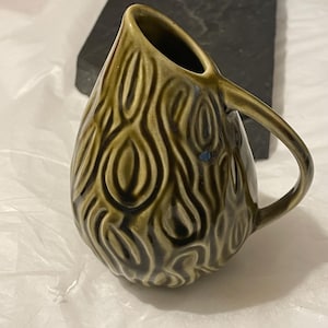 May include: A green ceramic jug with a textured, wood-grain design. The jug has a rounded body and a handle.