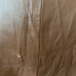 May include: Close-up of a brown leather surface with a subtle texture. The leather has a slight sheen and appears to be smooth and supple.