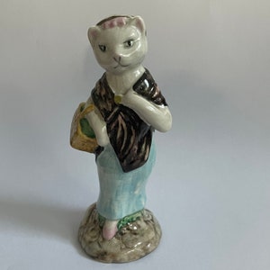 Può includere: Una statuetta in ceramica bianca di un gatto che indossa una giacca marrone e nera e una gonna blu. Il gatto tiene un cestino nella sua zampa e sta in piedi su una base verde e marrone.