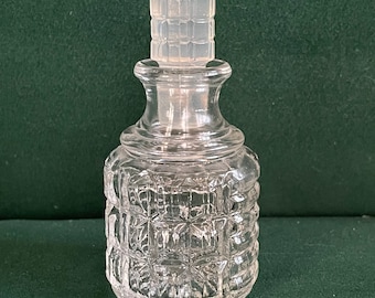 May include: A clear glass perfume bottle with a faceted stopper. The bottle has a rounded base and a textured, geometric pattern. The stopper is a clear, rectangular shape. The bottle is likely vintage and is displayed on a green surface.