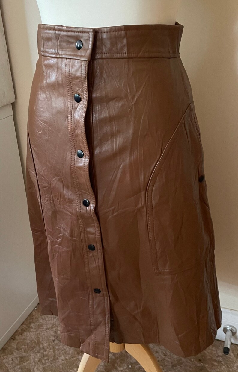 May include: A brown faux leather skirt with a button-up front and a side pocket. The skirt is knee-length and has a fitted waist.