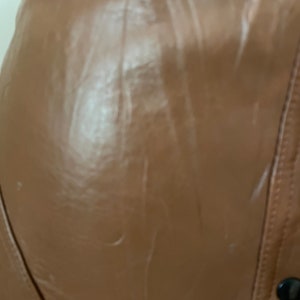 May include: A close-up of a brown leather jacket with a zipper and a button closure. The jacket has a smooth, shiny finish.