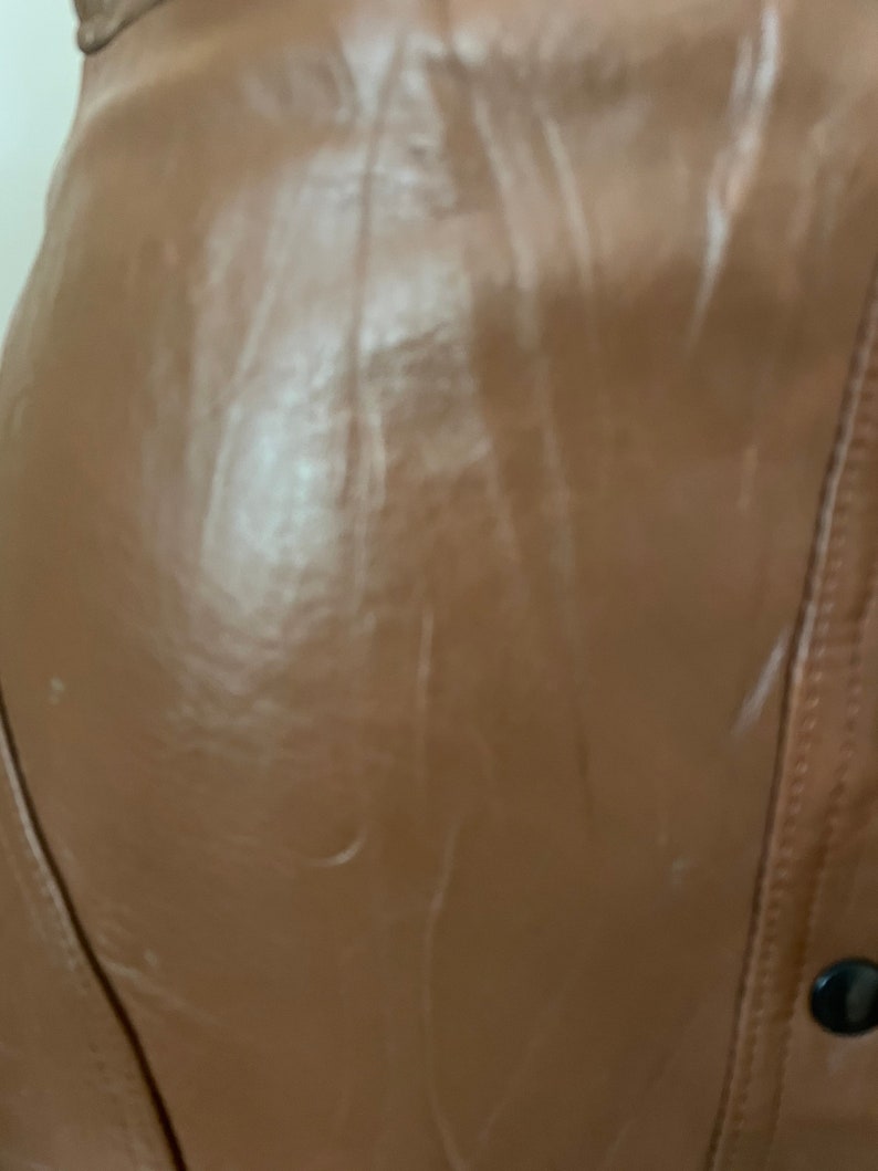 May include: A close-up of a brown leather jacket with a zipper and a button closure. The jacket has a smooth, shiny finish.