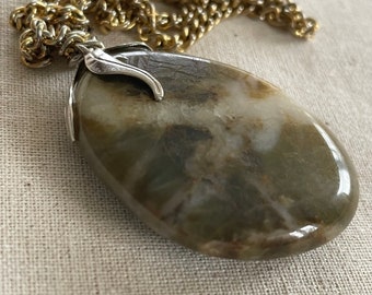 May include: A gold chain necklace with a large, oval-shaped pendant made of a light brown and white stone. The pendant has a silver-toned, leaf-shaped bail.