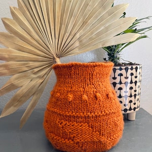 May include: An orange knitted vase with a textured design. The vase has a wide opening and a rounded body. The knitted pattern includes raised bobbles and a textured band near the base. A dried palm leaf and potted plants are in the background.