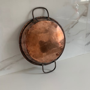 May include: A vintage copper pan with two iron handles. The pan is round and has a shiny copper finish.
