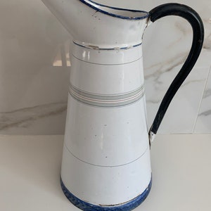 May include: White enamel pitcher with a black handle and blue trim. The pitcher has a tall, conical shape and is decorated with blue and gray stripes.
