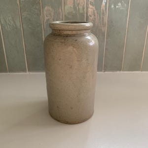 Può includere: Un barattolo cilindrico in gres con una finitura liscia color beige chiaro e un bordo arrotondato leggermente più scuro. La superficie presenta sottili macchioline naturali su tutto il corpo, conferendogli un aspetto strutturato e una forma semplice.