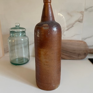 May include: A tall, brown stoneware bottle with a narrow neck and a cylindrical body. The bottle has a rustic, aged appearance, with variations in the brown color. A clear glass jar with a lid and a wooden cutting board are also visible.