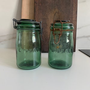 Peut inclure: Deux bocaux en verre vert avec des couvercles en métal et des fermetures à fil. Les bocaux ont le mot "LIDEALE" en relief sur le devant.