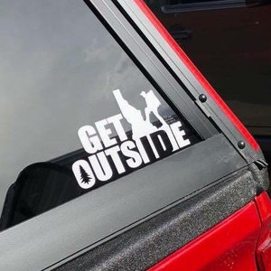May include: White vinyl decal with the text "GET OUTSIDE" and a silhouette of a person hiking. The decal is on the back window of a red truck.