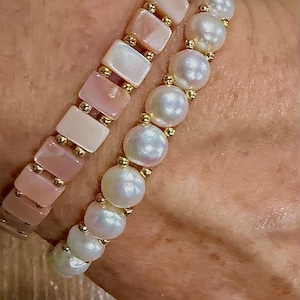 Puede incluir: Se muestran dos pulseras. Una tiene cuentas rosas rectangulares con detalles dorados. La otra pulsera tiene perlas blancas y redondas con detalles dorados. Ambas pulseras están en una muñeca.