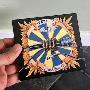 Op de afbeelding: Een vierkante zwarte cd-hoes met een microfoon die wordt vastgehouden door een skelet hand. Het ontwerp bevat een blauw en geel radiaal patroon, tarwearen en de tekst "Jet P" en "Losse Eindjes".