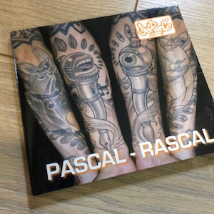 Op de afbeelding: Een boekomslag met een zwart-wit afbeelding van een arm met een tatoeage van een microfoon, een oog en een pen. De titel van het boek is "PASCAL - RASCAL" en het logo "Jeff PenCo" staat rechtsboven.