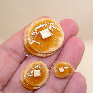 May include: Three miniature pancake stacks of varying sizes, each featuring a golden-brown surface topped with a glossy amber syrup glaze and a small, square, off-white butter pat centered on the top pancake of every individual stack.