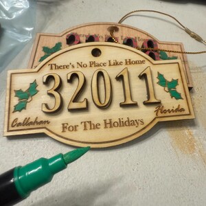 Op de afbeelding: Houten kerstornament met de tekst "There's No Place Like Home" en "32011 Callahan Florida For The Holidays". Het ornament heeft een hulstontwerp en een gouden touwtje om op te hangen. Een groene stift staat op de voorgrond.
